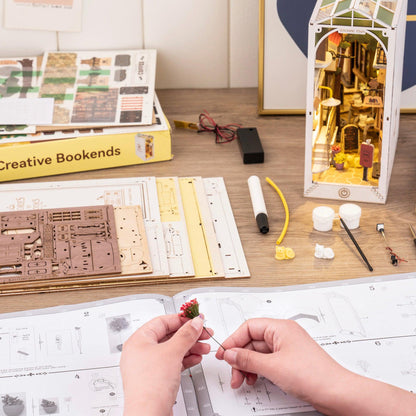 Hands assembling miniature Sunshine Town book nook with laser-cut wooden parts and instruction book.