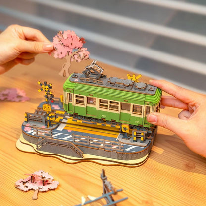 Hands positioning cherry blossom tree beside green tram model on Rolife Sakura Journey base.