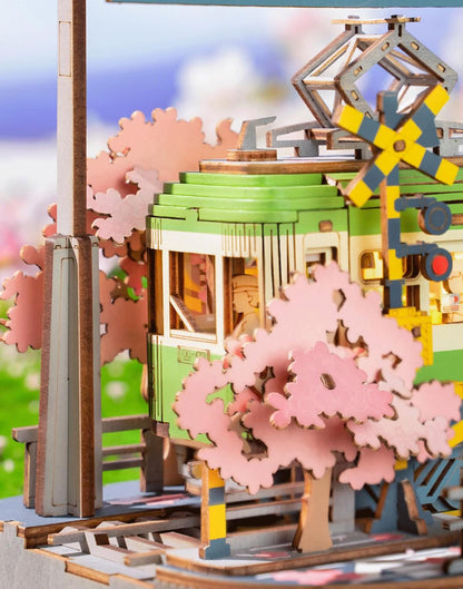 Close-up of yellow and black rail crossing signals beside green miniature tram and pink blossom branches.