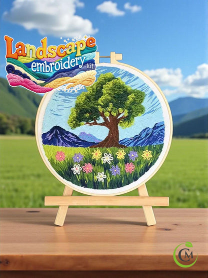 Landscape embroidery hoop with large green tree and wildflowers against hills and blue sky.